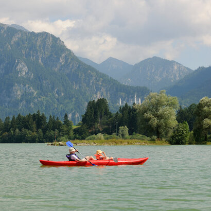 Das Bild fängt eine ruhige Szene mit zwei Personen ein, die Kajak fahren auf einem See. Die Kajakfahrer, mit Rettungswesten ausgestattet, paddeln in einem roten Kajak. Der See ist von majestätischen Bergen umgeben, was der Szene ein Abenteuergefühl verleiht. Der Himmel über ihnen ist ein klarer Blau, was auf einen schönen Tag für Outdoor-Aktivitäten hinweist. Die Gesamtstimmung des Bildes ist eine von Ruhe und dem Genießen der Natur.