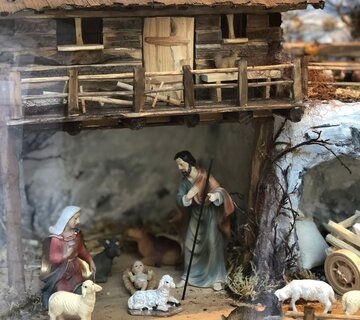 Das Bild zeigt eine Miniatur-Krippenszene, die die Geburt Jesu darstellt. Die Szene ist in einem Stall angesiedelt, welcher ein kleines hölzernes Gebäude mit Strohdach ist. Der Stall ist von einem hölzernen Zaun umgeben und im Hintergrund sind kleine Tiere, darunter Schafe und eine Kuh, zu sehen.  Im Vordergrund sind drei Figuren zu sehen: Maria, Josef und das Baby Jesus. Maria kniet nieder und hält das Baby Jesus in ihren Armen. Josef steht neben ihr und das Baby Jesus liegt in einer Futterkrippe.  Die Szene ist detailliert und bunt, mit verschiedenen Elementen wie Heu, Stroh und kleine Steine, die um den Stall herum verstreut sind. Die Gesamtkomposition des Bildes suggeriert eine friedliche und ruhige Atmosphäre und fängt so das Wesen der Weihnachtsgeschichte ein.