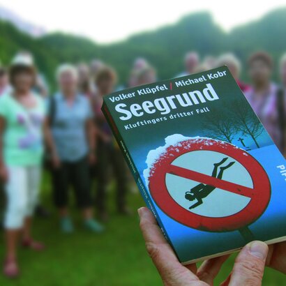 Das Bild zeigt eine Hand im Vorgergrund, die das Buch des Krimiromans "Seegrund" hält. Im Hintergrund ist eine kleine Menschengruppe zu sehen die vor dem Ufer des Alatsees steht und gebannt zusehen. 