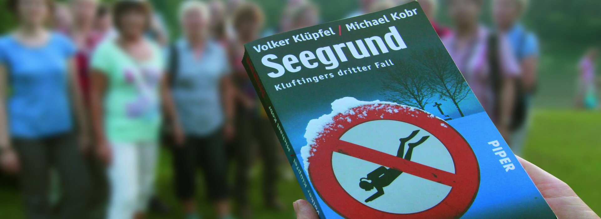 Das Bild zeigt eine Hand im Vorgergrund, die das Buch des Krimiromans "Seegrund" hält. Im Hintergrund ist eine kleine Menschengruppe zu sehen die vor dem Ufer des Alatsees steht und gebannt zusehen. 