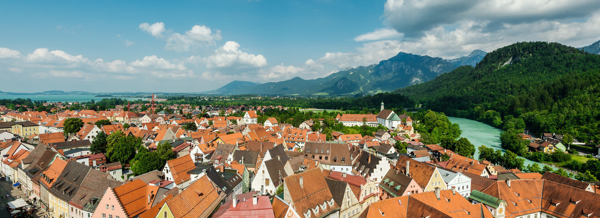 Das Bild zeigt eine Luftaufnahme der Stadt Füssen. Man sieht eine Vielzahl von sich aneinanderreihenden Dächern der Altstadt. Die Farben der größtenteils spitzen Giebeldächer reichen von hellem rot bis hin zu dunklem grau. Die Fassaden der Häuser sind größtenteils weiß, aber auch bunte Fassaden in Blau-, Gelb- oder Grüntönen sind zu sehen. An der Stadt entlang schlängelt sich das türkise Wasser des Flusses Lech. Im Hintergrund erheben sich majestätische Berge gegen den Himmel, deren Spitzen scheinbar die Wolken berühren. Der Himmel über ist Stadt ist blau mit der ein oder andern flauschig weißen Wolke. Das Wetter ist sonnig.