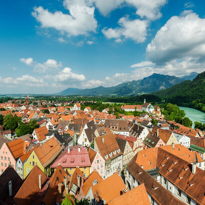 Das Bild zeigt eine Luftaufnahme der Stadt Füssen. Man sieht eine Vielzahl von sich aneinanderreihenden Dächern der Altstadt. Die Farben der größtenteils spitzen Giebeldächer reichen von hellem rot bis hin zu dunklem grau. Die Fassaden der Häuser sind größtenteils weiß, aber auch bunte Fassaden in Blau-, Gelb- oder Grüntönen sind zu sehen. An der Stadt entlang schlängelt sich das türkise Wasser des Flusses Lech. Im Hintergrund erheben sich majestätische Berge gegen den Himmel, deren Spitzen scheinbar die Wolken berühren. Der Himmel über ist Stadt ist blau mit der ein oder andern flauschig weißen Wolke. Das Wetter ist sonnig.