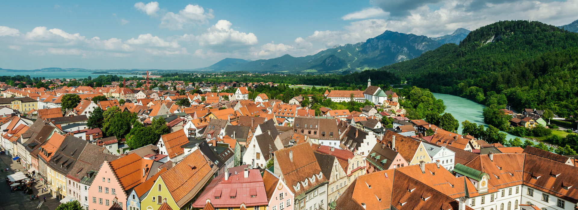 Das Bild zeigt eine Luftaufnahme der Stadt Füssen. Man sieht eine Vielzahl von sich aneinanderreihenden Dächern der Altstadt. Die Farben der größtenteils spitzen Giebeldächer reichen von hellem rot bis hin zu dunklem grau. Die Fassaden der Häuser sind größtenteils weiß, aber auch bunte Fassaden in Blau-, Gelb- oder Grüntönen sind zu sehen. An der Stadt entlang schlängelt sich das türkise Wasser des Flusses Lech. Im Hintergrund erheben sich majestätische Berge gegen den Himmel, deren Spitzen scheinbar die Wolken berühren. Der Himmel über ist Stadt ist blau mit der ein oder andern flauschig weißen Wolke. Das Wetter ist sonnig.
