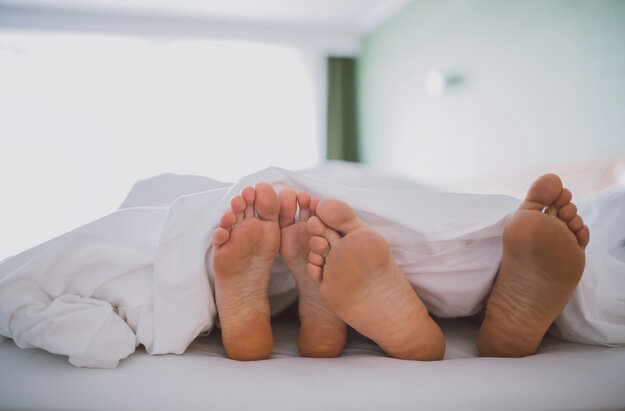 Das Bild zeigt zwei nackte Fußpaare, die unter einer weißen Decke hervorschauen. Die Füße sind so positioniert, dass sie fast verborgen sind. Die Decke, die das Hauptmotiv des Bildes ist, ist ordentlich verstaut, was eine behagliche und einladende Atmosphäre schafft. Die Kulisse ist verschwommen, was die Hauptmotive - die Füße und die Decke - betont. Die Gesamtzusammenstellung des Bildes suggeriert einen Moment der Entspannung und des Wohlbehagens