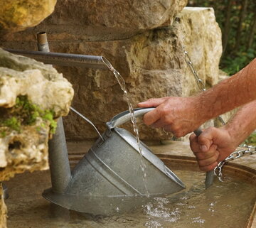 Das Bild zeigt die Nahaufnahme einer Metallgießkanne, die in ein Steinbecken mit Wasser gehalten wird. Aus einem Wasserhahn läuft Wasser herunter in das Becken. Das Becken ist eingerahmt von großen Natursteinen.
