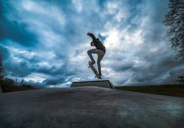  Das Bild fängt eine spannende Szene in einem Vergnügungspark ein. Der Fokus liegt auf einer großen, bunten Struktur, die einer Kletterwand ähnelt. Diese Struktur besteht aus verschiedenen Holzpfosten und Plattformen, die durch eine Vielzahl von Seilen und Drähten miteinander verbunden sind.  Mehrere Menschen sind dabei zu sehen, wie sie mit dieser Struktur interagieren, einige von ihnen klettern die Pfosten hinauf, während andere von den Plattformen schwingen. Die Struktur ist nicht nur ein Spielplatz, sondern auch ein Ort für Abenteuer.  Im Vordergrund gibt es eine kleine, hölzerne Plattform mit einer Brüstung. Diese Plattform ist mit einer Statue eines Ziegenbocks verziert, was der Szene eine Prise Fantasie verleiht.  Der Himmel darüber ist ein klares Blau, durchzogen von weißen Wolken, und bildet einen wunderschönen Hintergrund für die bunte Struktur. Das Bild ist eine lebendige Darstellung von Spaß und Abenteuer in einem Vergnügungspark.