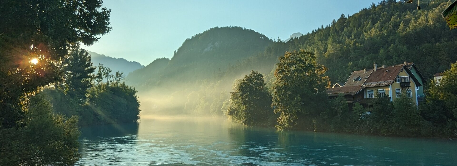 Das Bild fängt die ersten Sonnenstrahlen der Morgendämmerung am Lech ein, der sich dominant durch die Szene schlängelt. Der Fluss, ein tiefes Blau, liegt eingebettet in üppiges Grün, mit Bäumen und Büschen, die die Ufer säumen. Die ruhige Oberfläche des Flusses spiegelt die umliegende Landschaft wider. Rechts im Bild ist ein idyllisches Haus mit rotem Dach zu sehen. Obendrauf ist der Himmel ein helles Blau, durchzogen von ein paar Wolken. 