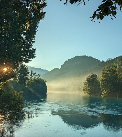 Das Bild fängt die ersten Sonnenstrahlen der Morgendämmerung am Lech ein, der sich dominant durch die Szene schlängelt. Der Fluss, ein tiefes Blau, liegt eingebettet in üppiges Grün, mit Bäumen und Büschen, die die Ufer säumen. Die ruhige Oberfläche des Flusses spiegelt die umliegende Landschaft wider. Rechts im Bild ist ein idyllisches Haus mit rotem Dach zu sehen. Obendrauf ist der Himmel ein helles Blau, durchzogen von ein paar Wolken. 