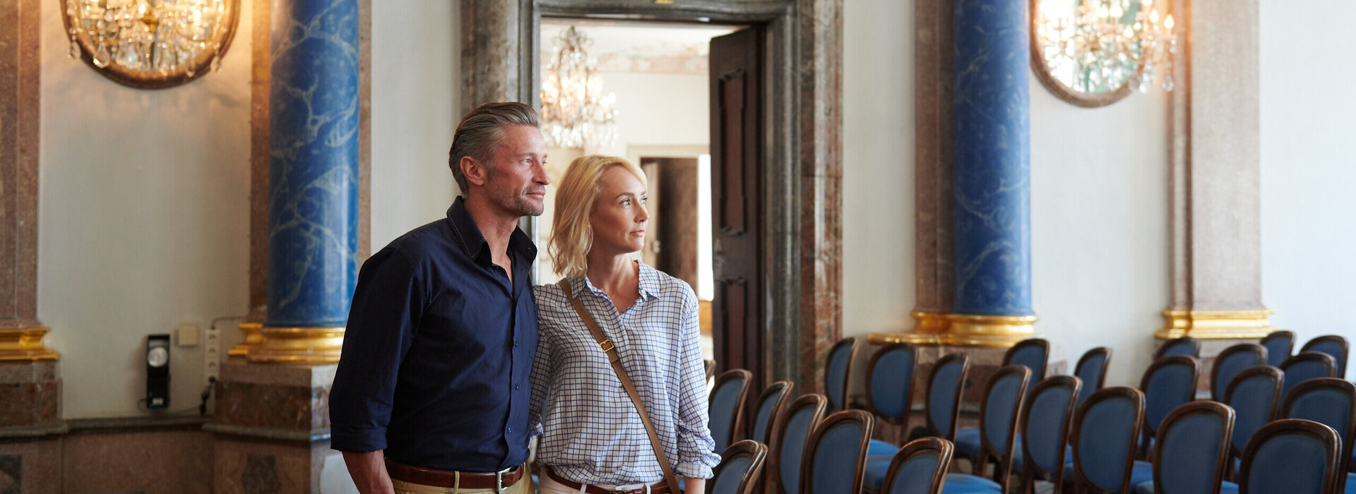 Das Bild zeigt einen Mann und eine Frau, die vor einem großen, aufwendig gestalteten Eingang stehen. Der Mann trägt ein blaues Hemd und Khakihosen, während die Frau ein weißes Oberteil und Jeans blau trägt. Sie blicken beide zur Kamera und die Frau hat ihren Arm um die Taille des Mannes gelegt. Der Raum, in dem sie sich befinden, ist geräumig und verfügt über blaue und goldene Akzente, mit mehreren aneinandergereihten Stühlen. Die Decke ist mit Kronleuchtern geschmückt und die Wände sind mit Gemälden dekoriert. Die Atmosphäre des Raumes wirkt edel und anspruchsvoll.