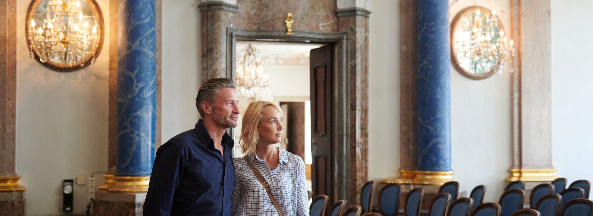 Das Bild zeigt einen Mann und eine Frau, die vor einem großen, aufwendig gestalteten Eingang stehen. Der Mann trägt ein blaues Hemd und Khakihosen, während die Frau ein weißes Oberteil und Jeans blau trägt. Sie blicken beide zur Kamera und die Frau hat ihren Arm um die Taille des Mannes gelegt. Der Raum, in dem sie sich befinden, ist geräumig und verfügt über blaue und goldene Akzente, mit mehreren aneinandergereihten Stühlen. Die Decke ist mit Kronleuchtern geschmückt und die Wände sind mit Gemälden dekoriert. Die Atmosphäre des Raumes wirkt edel und anspruchsvoll.
