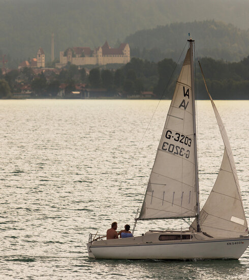 Das Bild zeigt im Vordergrund ein weißes Segelboot auf einem See fahren. Es hat die Segel gehisst. Auf dem Boot sitzen zwei Personen. Im Hintergrund erhebt sich das Hohe Schloss über der Stadt Füssen. Das Ufer ist mit hohen Bäumen bewachsen. Im Hintergrund erheben sich bewaldete Berge. Die Abendsonne taucht die Szene in ein warmes Licht. 