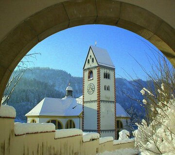 Das Bild zeigt die Kirche St. Mang in Füssen in einer winterlichen Ansicht. Die Kirche erkennt man im Hintergrund eines durch schneebedeckten Tores des Hohen Schlosses. 