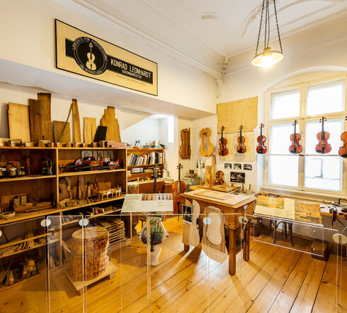 Man blickt in eine Geigenbauerwerkstatt. Viele Geigen und Bratschen hängen an einer Schnur nebeneinander. Ein Regal ist mit Hölzern, Schablonen und Latten gefüllt. Ein Regal mit Gläsern und Flaschen voller Flüssigkeiten. Vor einem Fenster steht eine Werkbank. Über der Szenerie hängt ein Firmenschild. Darauf steht: „Konrad Leonhart, Geigenbaumeister.“ Am Schreibtisch im Hintergrund lehnt ein Cello. Das Regal über dem Schreibtisch ist mit Büchern und Ordnern gefüllt. 