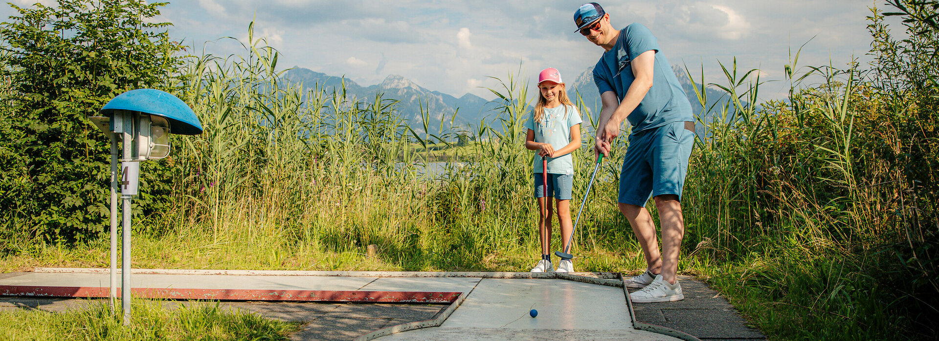 Das Bild zeigt eine friedliche Szene eines Mannes und eines jungen Mädchens, die Golf spielen auf einer grünen Wiese. Der Mann, in einem blauen Hemd und blauen Shorts gekleidet, ist dabei, seinen Golfschläger zu schwingen und auf den Golfball zu zielen, der auf dem Boden liegt. Das Mädchen, das einen pinken Hut trägt, steht in der Nähe und beobachtet aufmerksam den Schwung des Mannes. Die Wiese, auf der sie sich befinden, ist üppig und grün, mit hohen Gräsern, die sanft im Wind wehen. In der Ferne erstreckt sich ein majestätisches Gebirge über den Horizont und verleiht der Szene eine beruhigende Ruhe. Der Himmel ist ein klarer blauer Farbton, mit flauschigen weißen Wolken verstreut. Das Bild vermittelt ein Gefühl von Leichtigkeit und Freude an der Natur.