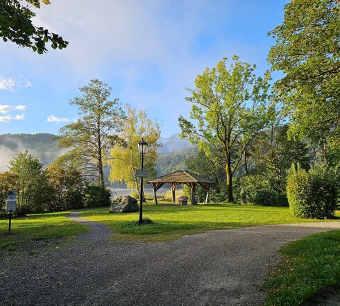 Vom Betrachter führen zwei Kieswege weg. Einer führt nach rechts außerhalb des Blickfelds, der andere geradeaus auf einen hölzernen Pavillon zu, der auf einer Wiese steht. Um den Pavillon herum stehen grüne Laubbäume und Büsche. Im Hintergrund erheben sich bewaldete Hügel und Berge. Der Himmel ist blau. Über dem Wasser des Sees, liegt noch der Morgennebel. Die Sonne scheint.    