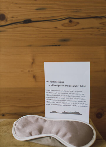 Das Bild zeigt eine beige Augen-Schlafmaske und ein weißes Blatt Papier, das dahintersteht. Die Überschrift auf dem Papier lautet „Wir kümmern und um Ihren guten und gesunden Schlaf“. Darunter ist weiterer Text platziert. Im Hintergrund ist ein Holzwand. 