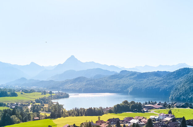 Das Bild den Ort Weißensee von einer Anhöhe aus. Man sieht den gleichnamigen See. Dahinter erheben sich bewaldete Hügel und unzählige Gipfel. Der Himmel ist blau. Die Sonne scheint.  