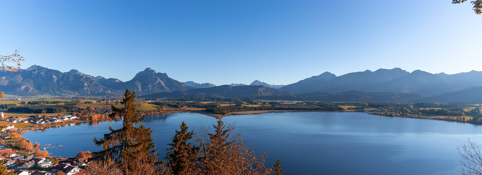 Das Herbstfoto zeigt die Aussicht auf den Hopfensee und die Alpengipfel rund um Füssen im Allgäu von der Burgruine Hopfen aus.