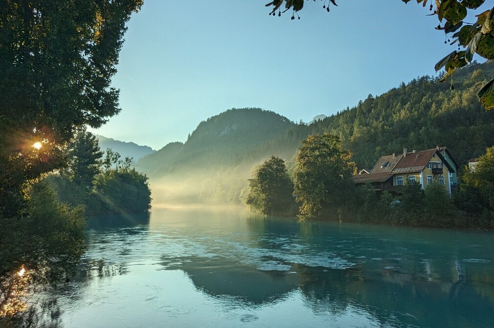 Das Bild zeigt eine ruhige Landschaft, die von einem friedlichen Fluss beherrscht wird, der sich durch die Szene schlängelt. Der türkise Fluss Lech fließt friedlich dahin, eingebettet in üppige Vegetation, mit Bäumen und Sträuchern, die die Ufer säumen. Die ruhige Oberfläche des Flusses spiegelt die umgebende Landschaft wider. Auf der rechten Flussseite steht ein charmantes gelbes Haus mit rotem Dach. Der klare und blaue Morgenhimmel ist wolkenlos. Die Sonne ist gerade aufgegangen. Ihre Strahlen bahnen sich ihren Weg durch die Äste der Bäume, die am Ufer stehen. 