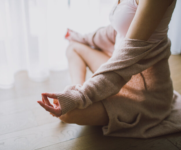 Das Bild zeigt eine Frau, die im Schneidersitz auf dem Holzboden sitzt. Sie sitz vor einem hellen Fenster, das mit einem bodenlangen Stohr verhängt ist. Sie eine beige Strickjacke, die ihr von den Schultern gerutscht ist, ein rosafarbenes Tanktop und eine kurze Hose. Ihre Hände ruhen auf ihren Knien. Mittelfinger und Daumen beider Hände berühren sich.     