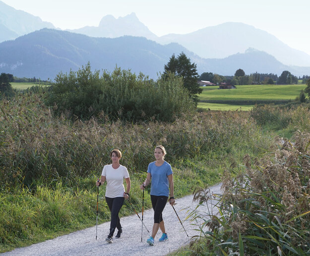 Das Bild zeigt zwei Frauen, die mit Sportkleidung und Nordic Walking stecken einen Wanderweg entlang laufen, der von Schilf flankiert ist. Sie laufen auf den Betrachter zu. Im Hintergrund sind grüne Wiesen und hohe Berge zu sehen. Der Himmel ist blau. Die Sonne scheint.   
