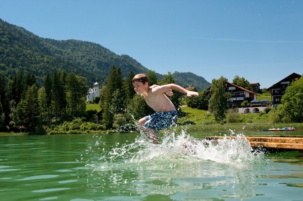 Das Bild zeigt einen Jungen, der in den Weißensee springt. Er hat die Wasseroberfläche bereits durchbrochen und das Wasser spritzt in alle Richtungen umher. Der Junge trägt blaue Badehosen. Der Weißensee ist von grünen Bäumen und Hügeln umgeben. Der Himmel ist blau. Die Sonne scheint.  