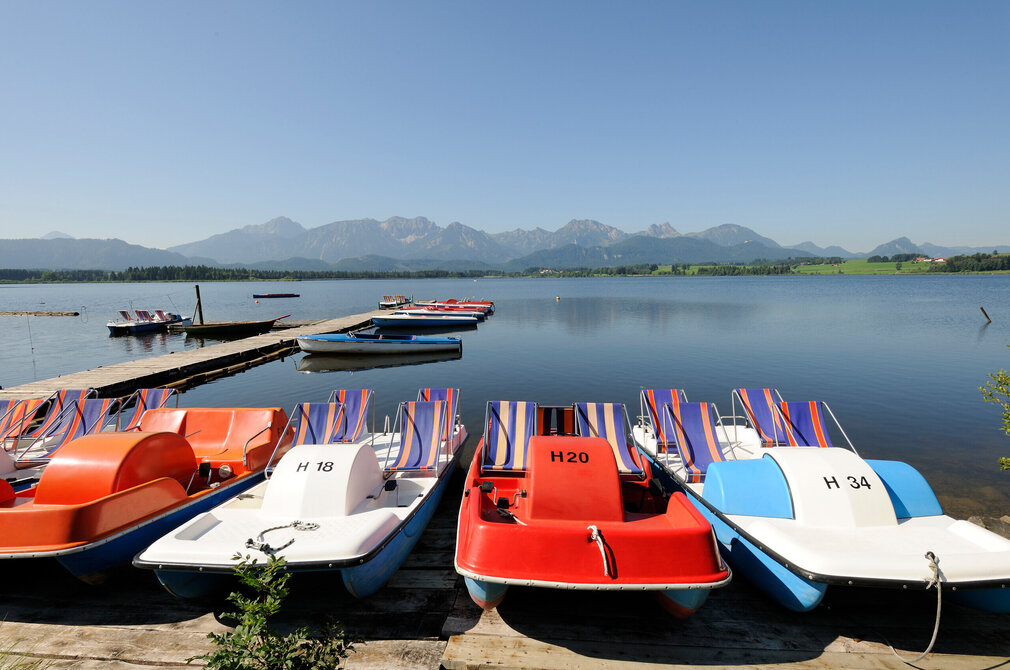 Das Bild fängt eine ruhige Szenerie an einem Steg ein, an den eine Reihe bunter Tretboote aufgereiht sind. Die Boote, die in lebhaften Farben wie Rot, Blau und Orange gestrichen sind, liegen in einem ruhigen Gewässer – dem Hopfensee. Der Steg ist aus Holz gefertigt. Im Hintergrund erheben sich majestätische Berge. Der Himmel ist blau und klar. Das Bild strahlt eine Atmosphäre der Ruhe und Erholung aus und lädt dazu ein, sich die Beobachter auf einem dieser Boote zu stellen und einen friedlichen Tag auf dem Wasser zu genießen.