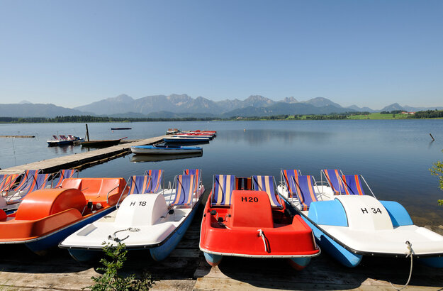 Das Bild fängt eine ruhige Szenerie an einem Steg ein, an den eine Reihe bunter Tretboote aufgereiht sind. Die Boote, die in lebhaften Farben wie Rot, Blau und Orange gestrichen sind, liegen in einem ruhigen Gewässer – dem Hopfensee. Der Steg ist aus Holz gefertigt. Im Hintergrund erheben sich majestätische Berge. Der Himmel ist blau und klar. Das Bild strahlt eine Atmosphäre der Ruhe und Erholung aus und lädt dazu ein, sich die Beobachter auf einem dieser Boote zu stellen und einen friedlichen Tag auf dem Wasser zu genießen.