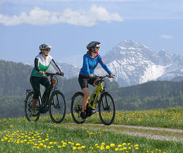 Das Bild zeigt zwei Frauen beim Fahrradfahren. Beide Frauen tragen Sportklamotten, Fahrradhosen und Helme. Beide lächeln. Sie fahren auf einem unbefestigten Feldweg, der durch eine Wiese führt. Die Wiese ist übersäht von unzähligen gelben Löwenzahlblüten. Im Hintergrund erheben sich schneebedeckte Berge und grün, bewaldete Hügel. Der Himmel ist blau. Die Sonne scheint. 