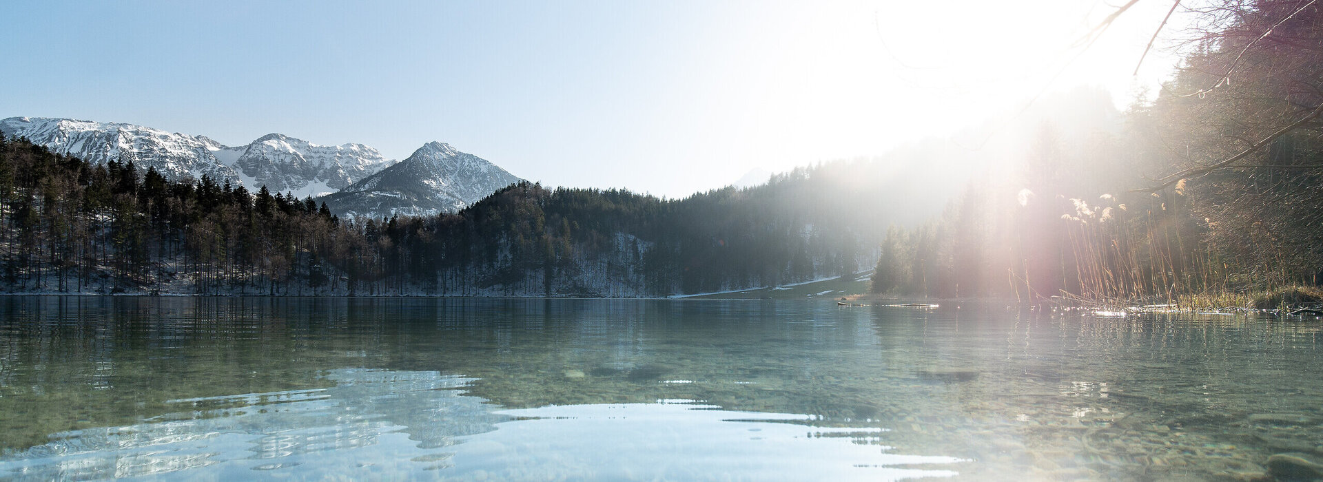 Das Bild fängt eine harmonische Szene eines in einem gebirgigen Gebiet gelegenen Sees ein – des Alatsees. Der See, mit seiner ruhigen und friedlichen Oberfläche, reflektiert die umgebende Landschaft. Die Sonne scheint hell, wirft ein warmes Licht auf das Wasser und erzeugt einen schönen Kontrast zu den dunkleren Farben der Berge. Die Berge, mit Schnee bedeckt, erheben sich majestätisch im Hintergrund und verleihen der Szene ein Gefühl von Größe. Der Himmel über dem See ist ein klarer Blau, was das Gesamtbild des Bildes weiter verschönert. 