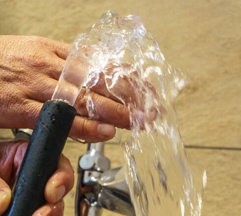 Das Bild zeigt zwei Hände die einen Wasserschlauch halten. Aus dem Wasserschlauch kommt ein klares Wasser, das über die eine Hand läuft. Im Hintergrund ist ein silberner Wasserhahn zu sehen.
