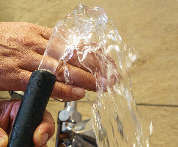 Das Bild zeigt zwei Hände die einen Wasserschlauch halten. Aus dem Wasserschlauch kommt ein klares Wasser, das über die eine Hand läuft. Im Hintergrund ist ein silberner Wasserhahn zu sehen.      