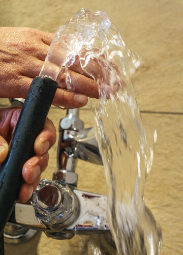Das Bild zeigt zwei Hände die einen Wasserschlauch halten. Aus dem Wasserschlauch kommt ein klares Wasser, das über die eine Hand läuft. Im Hintergrund ist ein silberner Wasserhahn zu sehen.      