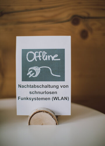 Das Bild zeigt einen Blatt Papier, das in einer Holzhalterung steht. Auf dem Papier ist zu lesen „Nachtabschaltung von schnurlosen Funksystemen (WLAN). Im Hintergrund ist eine einfache Holzwand zu sehen. 