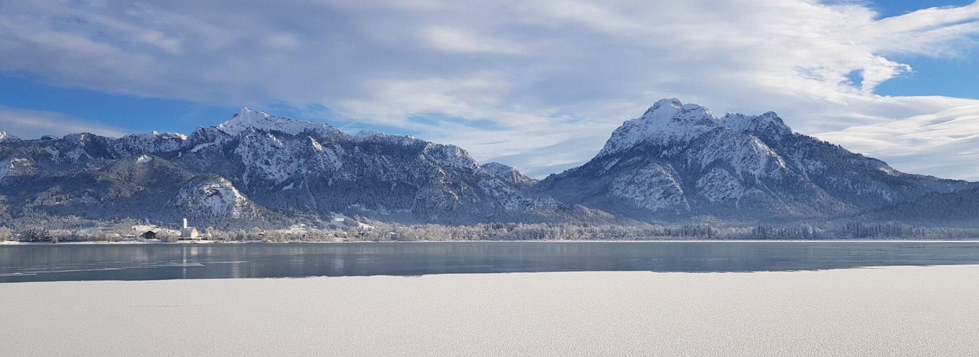 Das Bild zeigt ein atemberaubendes Panorama eines schneebedeckten Gebirgszuges unter einem klaren blauen Himmel. Die Berge, mit ihren zackigen Gipfeln, erheben sich majestätisch im Hintergrund. Die Vordergrunddomäne wird von einem stillen See dominiert, dessen ruhiges Wasser die Berge widerspiegelt. Die Oberfläche des Sees ist unberührt, was zur Ruhe des Bildes beiträgt. Die Gesamtkomposition des Bildes suggeriert eine friedliche und ruhige Landschaft, unberührt von menschlichem Eingriff. 