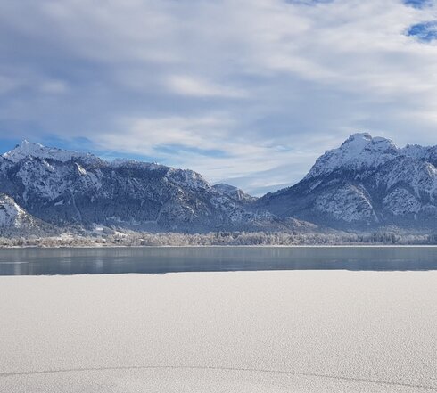 Das Bild zeigt ein atemberaubendes Panorama eines schneebedeckten Gebirgszuges unter einem klaren blauen Himmel. Die Berge, mit ihren zackigen Gipfeln, erheben sich majestätisch im Hintergrund. Die Vordergrunddomäne wird von einem stillen See dominiert, dessen ruhiges Wasser die Berge widerspiegelt. Die Oberfläche des Sees ist unberührt, was zur Ruhe des Bildes beiträgt. Die Gesamtkomposition des Bildes suggeriert eine friedliche und ruhige Landschaft, unberührt von menschlichem Eingriff. 