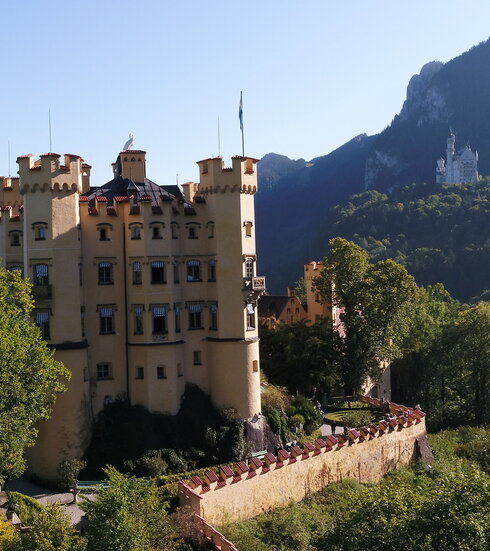 Das Bild zeigt im Vordergrund das gelbe Schloss Hohenschwangau. Es ist vier Stockwerke hoch und hat an jeder Ecke einen Turm. Um den Dachbereich verlaufen Zinnen. Auf dem Dach steht eine überlebensgroße, weiße Schwanenfigur. Auf einem der Türme ist eine Fahne zu sehen. Im Hintergrund ist das weiße Schloss Neuschwanstein zu sehen. Beide Schlösser sind von Wäldern umgeben. Beide Schlösser stehen auf Anhöhen vor einem Berg. Das Schloss Neuschwanstein steht ein wenig höher als das Schloss Neuschwanstein. Der Himmel ist blau. Die Sonne scheint. 