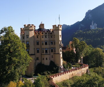 Das Bild zeigt im Vordergrund das gelbe Schloss Hohenschwangau. Es ist vier Stockwerke hoch und hat an jeder Ecke einen Turm. Um den Dachbereich verlaufen Zinnen. Auf dem Dach steht eine überlebensgroße, weiße Schwanenfigur. Auf einem der Türme ist eine Fahne zu sehen. Im Hintergrund ist das weiße Schloss Neuschwanstein zu sehen. Beide Schlösser sind von Wäldern umgeben. Beide Schlösser stehen auf Anhöhen vor einem Berg. Das Schloss Neuschwanstein steht ein wenig höher als das Schloss Neuschwanstein. Der Himmel ist blau. Die Sonne scheint. 