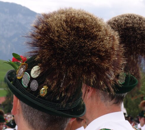 Das Bild zeigt zwei Herren mit weißen Hemden und grünen Lederhosenträgern. Auf Ihren Köpfen tragen sie grüne Trachtenhüte mit zahlreichen Abzeichen aus Metall. Dazu ist auf jedem Hut je ein ausladender Gamsbart befestigt. Die Männer drehen dem Betrachter den Rücken zu. Man sieht nur ihre Köpfe und einen Teil der Oberkörper. Im Hintergrund kann man weitere Personen in Tracht erkennen. Der Himmel ist wolkenverhangen.