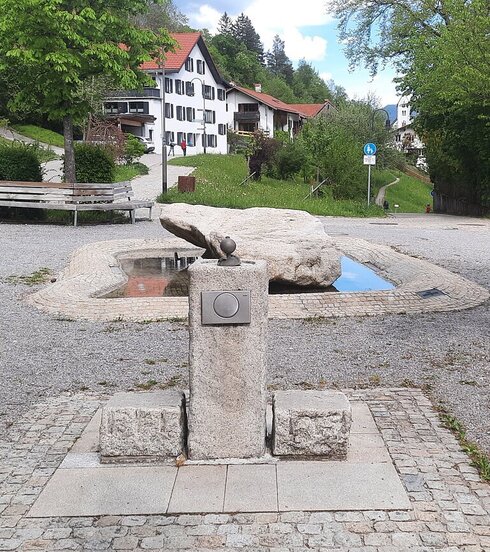 Das Bild zeigt eine friedliche Parkszene. Im Zentrum der Szene befindet sich ein großer grauer Stein, der als Hauptfokus dient. Dieser Stein ist von einer kreisrunden Steinpfad umgeben, die ein Gefühl von Einheit und Harmonie schafft. Die Pfad ist von üppigem grünem Gras begrenzt, was der Szene einen Hauch von Natur verleiht. Im Hintergrund sind mehrere weiße Gebäude zu sehen, deren Architektur mit den natürlichen Elementen im Vordergrund kontrastiert. Die Gebäude sind eingebettet zwischen Bäumen, was die friedliche Atmosphäre des Parks weiter verstärkt. Auf der rechten Seite des Bildes steht eine hölzerne Bank, die Besucher einlädt, sich hinzusetzen und die friedliche Umgebung zu genießen. Der rustikale Charme der Bank ergänzt den allgemeinen Stil des Parks. Der Himmel über ist ein klarer Blau, was auf einen hellen und sonnigen Tag hinweist. Das Bild fängt das Wesen eines gut gepflegten Parks ein, in dem Natur und Architektur in Harmonie leben.