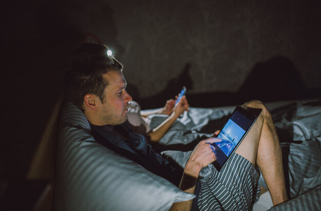 Das Bild zeigt zwei Personen, die im Bett liegen. Sie lehnen nebeneinander am Kopfende. Der Mann im Vordergrund sieht in ein Tablet. Die Person im Hintergrund hat ein Handy in der Hand. Es ist dunkel nur die Nachttischlampen erhellen das Zimmer.    