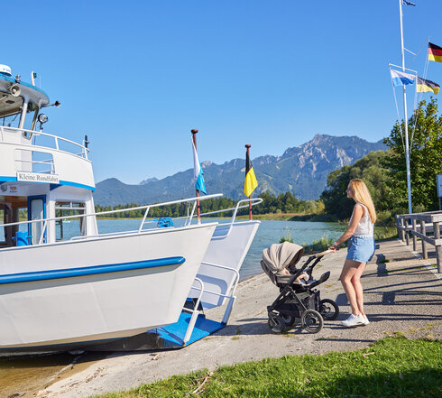 Das Bild zeigt eine beschauliche Szene am Steg. Ein großes weißes Boot liegt am Pier, seine blaue Streifen geben der ansonsten monochromen Erscheinung einen Farbakzent. Das Boot ist nicht allein, denn es wird von einigen Fahnen begleitet, die im Wind flattern.  Auf dem Steg genießt eine Familie ihre Zeit. Eine Frau, in einem blauen Kleid gekleidet, schiebt einen Kinderwagen, vielleicht bereitet sie sich auf einen Ausflug vor. Sie ist begleitet von einem Mann, der ein Kind in den Armen hält. Ihre Präsenz verleiht der Szene ein Gefühl von Wärme und familiärer Liebe.  Der Hintergrund des Bildes ist ein atemberaubender Gebirgszug, der einen faszinierenden Kontrast zu den ruhigen Gewässern des Sees bietet. Der klare blaue Himmel darüber bereichert die Schönheit der Szene zusätzlich.  Insgesamt malt das Bild ein Bild eines friedlichen Tages am Steg, mit der Familie, die ihre Zeit inmitten der natürlichen Schönheit des Sees und der Berge genießt.