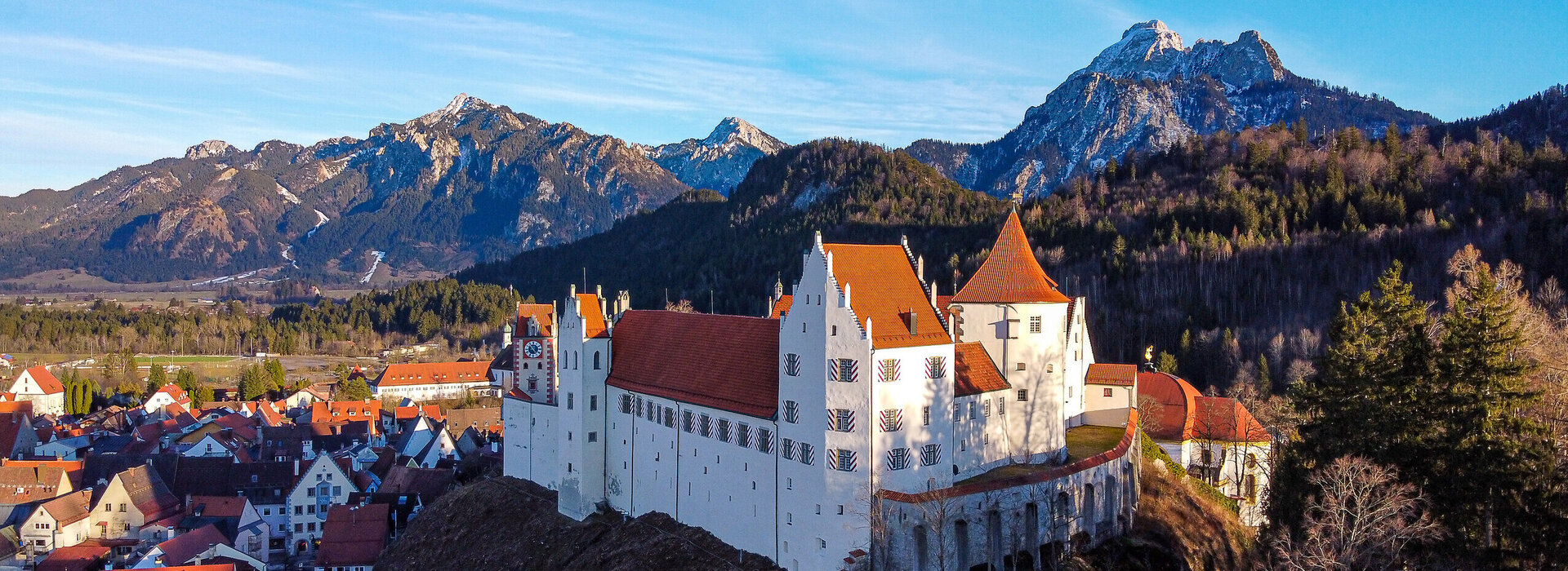 Man blickt von oben auf das Hohe Schloss, das auf einer Anhöhe über Füssen thront. Darunter sieht man die Häuser der Altstadt, die dicht aneinandergebaut wurden. Im Hintergrund erheben sich schneebedeckte Berge und bewaldete Hügel. Der Himmel ist blau. Die Sonne scheint. 