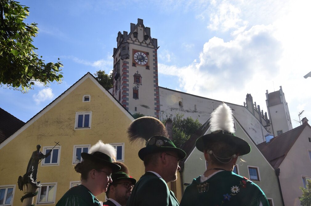 Das Bild zeigt mehrere Personen in Tracht. Man erkennt nur ihre Köpfe und Oberkörper. Die Männer tragen grüne Trachtenjacken. Die Frau mit Blüten verzierte Schultertücher. Alle tragen grüne Trachtenhüte. Die Männer haben Gamsbärte, die Frauen Adlerflaume an ihren Hüten befestigt. Im Hintergrund sind bunte Hausfassaden zu erkennen. Darüber thront der Uhrturm des Hohen Schlosses. Der Himmel ist blau. Die Sonne scheint.