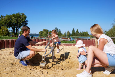  Das Bild zeigt eine herzerwärmende Szene am sandigen Strand. Ein Mann, der ein blaues Hemd und beigefarbene Shorts trägt, kniet neben einem kleinen Kind. Er hält ein metallisches Objekt, möglicherweise ein Spielzeug, in der Hand. Das Kind, das eine pinke Mütze trägt, steht neben ihm und hält sich an dem Bein des Mannes fest.  Etwas weiter entfernt sitzt eine Frau auf dem sandigen Strand. Sie trägt ein weißes Hemd und blaue Shorts. Ihr Blick ist auf den Mann und das Kind gerichtet, was auf eine enge Beziehung zwischen ihnen hinweist.  Der Hintergrund des Bildes zeigt eine idyllische Strandszene. Der Himmel ist klar und blau, und das Wasser erscheint ruhig. Die Gesamtstimmung des Bildes ist eine ruhige Entspannung und das Genießen eines Tages am Strand.