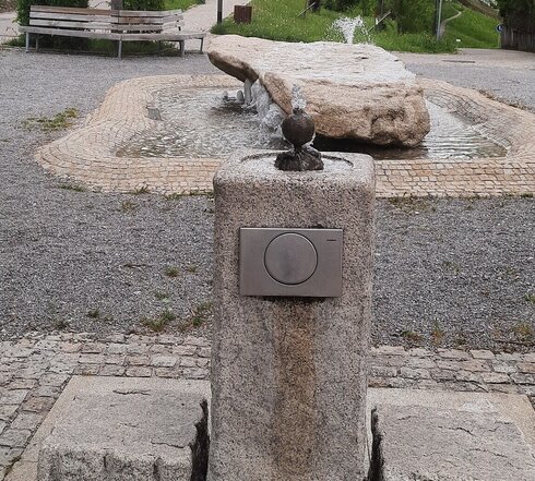 Das Bild zeigt im Vordergrund eine Steinsäule, neben der jeweils links und rechts zwei Steinquader liegen. Aus der Oberseite der Steinsäule sprudelt Wasser. An der, der Kamera zugewandten Seite der Steinsäule ist ein Schalter befestigt. Im Hintergrund liegt ein großer Steinblock, um den unterschiedlich hohe Wasserfontänen nach oben sprudeln. Nur eine Fontäne überragt den Steinblock. Der Steinblock liegt in einem mit Kopfsteinpflaster ausgekleideten Wasserbassin. Im Hintergrund sind Häuser, Bäume und eine Straße zu sehen.  