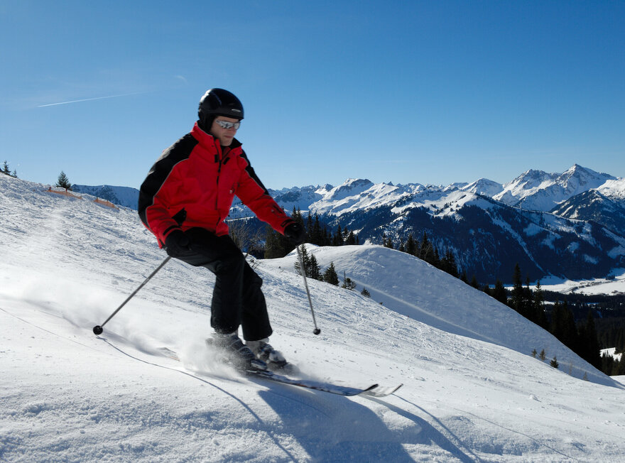 Das Bild zeigt eine Person in Skikleidung und Helm beim Skifahren auf einer ansonsten menschenleeren Piste.  Die Landschaft ringsum ist tief verschneit. Im Hintergrund erheben sich die schneebedeckten Berge. Zahlreiche Gipfel sind zu erkennen. Der Himmel ist blau. Die Sonne scheint. 