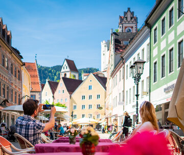 Das Foto zeigt einen Mann und eine Frau, die an einem Tisch in der Fußgängerzone von Füssen sitzen, wahrscheinlich vor einem Cafe. Die Straße ist links und rechts von Gebäuden gesäumt. Es ist Sommer. Die Sonne scheint. Über den Häusern erheben sich die Türme vom Hohen Schloss. Der Mann und die Frau haben Ihren Blick weg von der Kamera gerichtet. Der Mann hat ein Handy in der Hand und macht gerade ein Foto.