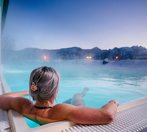 Das Bild zeigt eine Frau, die in Badekleidung in einem dampfenden Outdoor-Pool liegt. Sie dreht dem Betrachter den Rücken zu und hat die Haare hochgesteckt. Ihre Arme ruhen auf dem Poolrand. Die Landschaft im Hintergrund ist tief verschneit.  