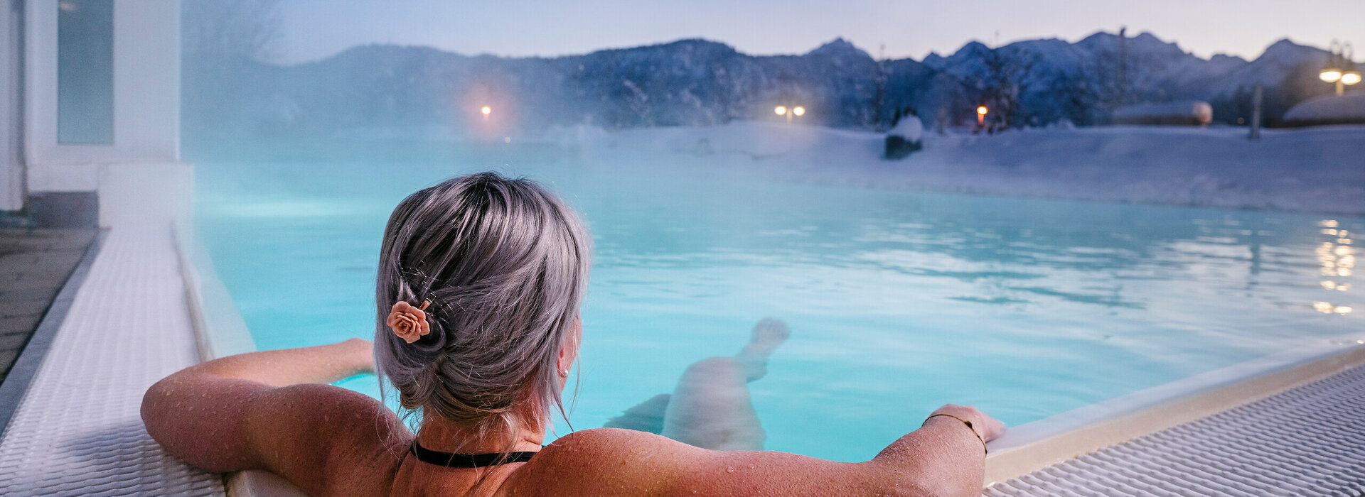 Das Bild zeigt eine Frau, die in Badekleidung in einem dampfenden Outdoor-Pool liegt. Sie dreht dem Betrachter den Rücken zu und hat die Haare hochgesteckt. Ihre Arme ruhen auf dem Poolrand. Die Landschaft im Hintergrund ist tief verschneit.  